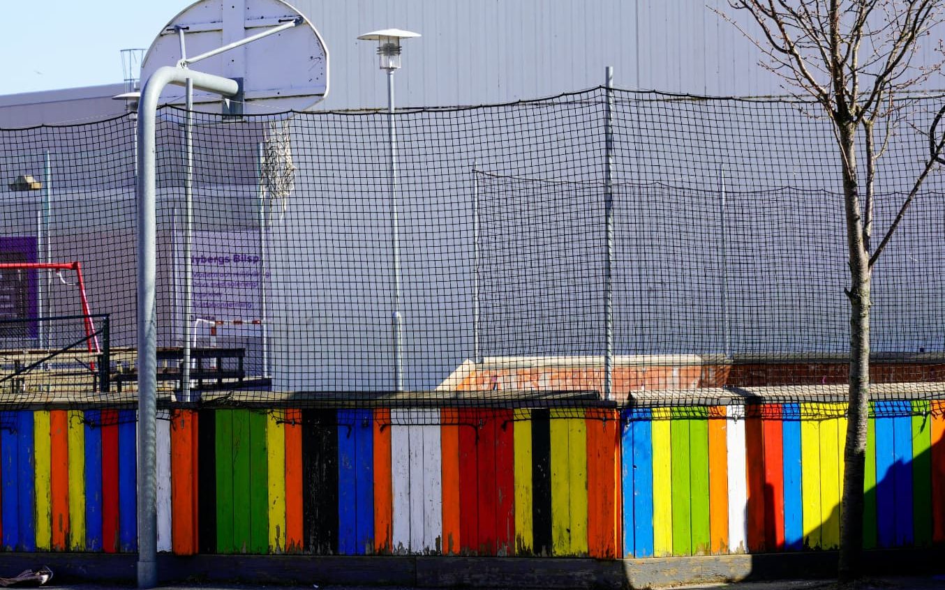 An empty school playground with a basketball hoop in front of a brightly painted wooden fence in red, yellow, green and blue