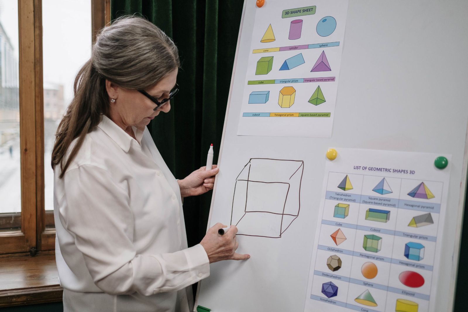 A teacher drawing a cube on a whiteboard beside printed sheets of 3D geometric shapes